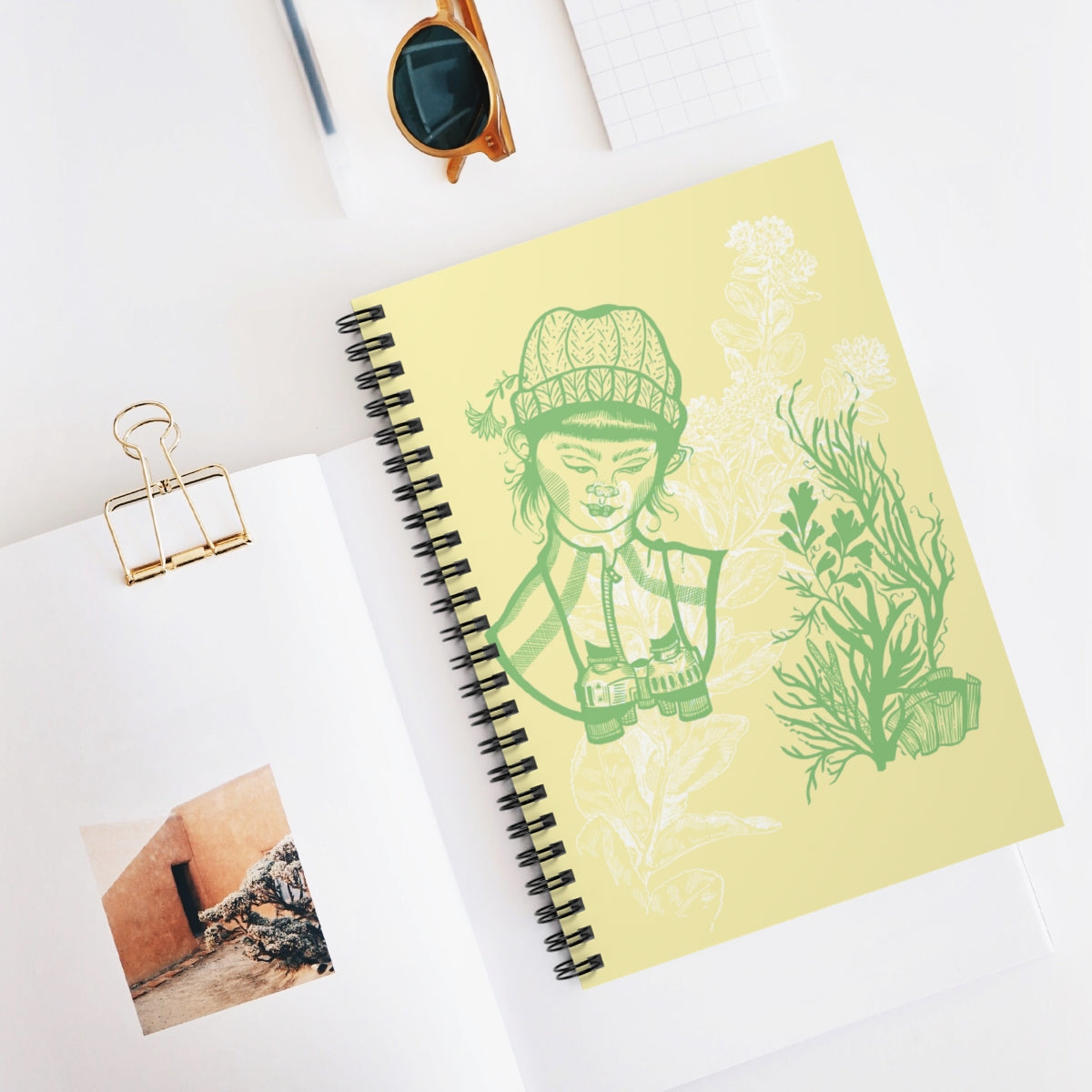 A female biologist with binoculars looks at plants sketched in green ink on a tan cover. The image is underlaid with a white flower watermark. The book is spiral bound and has lined pages. 