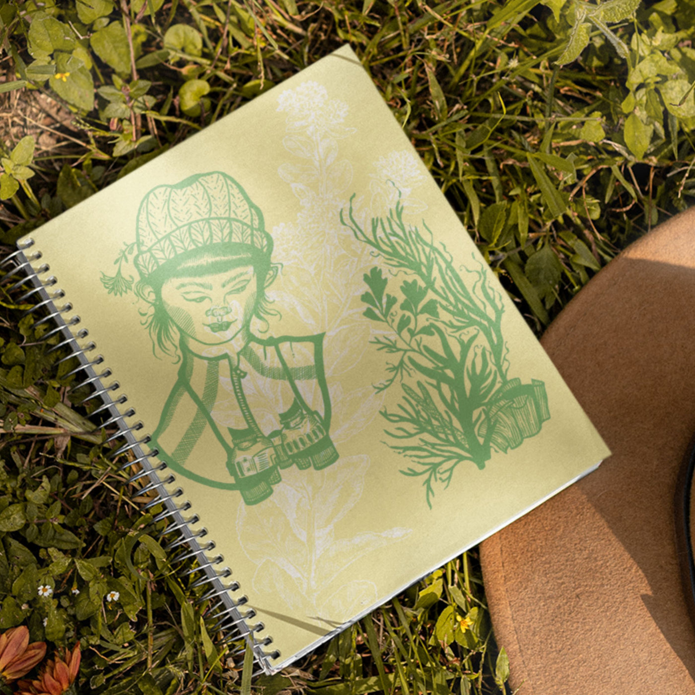 A female biologist with binoculars looks at plants sketched in green ink on a tan cover. The image is underlaid with a white flower watermark. The book is spiral bound. 